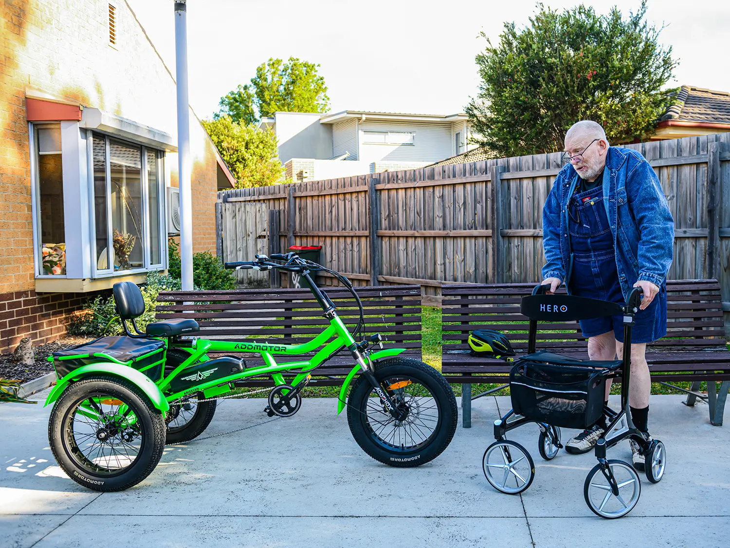 An older man using a walker stands near a bright green Addmotor M-360 electric tricycle parked beside a wooden bench. The tricycle features a comfortable padded seat with back support, fat tires, and a rear storage compartment, ideal for seniors and mobility assistance.